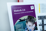 Das Bild zeigt ein Lehrmaterial namens "Elektrik 2.0", das sich mit den Themen Induktion und Wechselspannung beschäftigt. Zudem ist ein Junge abgebildet, der mit Experimentiergeräten arbeitet, um elektrische Phänomene zu erforschen.