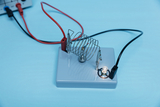 Das Bild zeigt eine experimentelle Vorrichtung mit einer spiralförmigen Feder und einer LED auf einer grauen Basis, die durch farbige Kabel verbunden ist. Diese Anordnung dient der Veranschaulichung von elektrischen Schaltungen und Experimenten im Bildungsbereich.