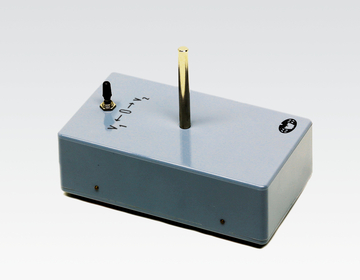 Das Bild zeigt ein Experimentiergerät mit einem Metallstift in der Mitte und zwei Anschlussbuchsen an der Seite. Es scheint für den Einsatz im Bildungsbereich entwickelt worden zu sein, vermutlich zur Durchführung physikalischer Experimente.