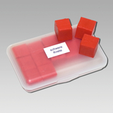 Das Bild zeigt eine transparente Box, die mit roten Würfeln gefüllt ist, die als "schwere Knete" beschriftet sind. Einige Würfel liegen außerhalb der Box, während die restlichen darin aufgereiht sind und ein praktisches Experimentiermaterial darstellen.
