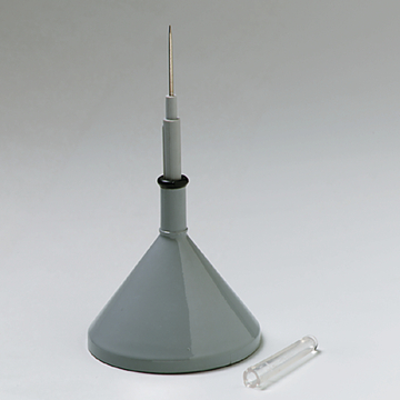 Das Bild zeigt einen grauen Trichter mit einer langen Spitze, der für wissenschaftliche Experimente verwendet wird. Neben dem Trichter liegt ein kleines, transparentes Röhrchen.