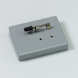 Das Bild zeigt ein graues experimentelles Gerät mit einem Schalter und zwei verfügbaren Anschlussbuchsen. Es dient vermutlich der Durchführung von elektrischen Experimenten im Bildungsbereich.