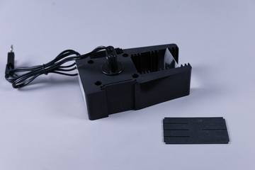 Das Bild zeigt ein schwarzes Gerät mit einem Motor und einem dazugehörigen Kabel, das möglicherweise für Versuche in der Schule verwendet wird. Neben dem Gerät liegt ein schwarzes, rechteckiges Teil, das vermutlich als Zubehör oder Unterstützung dient.