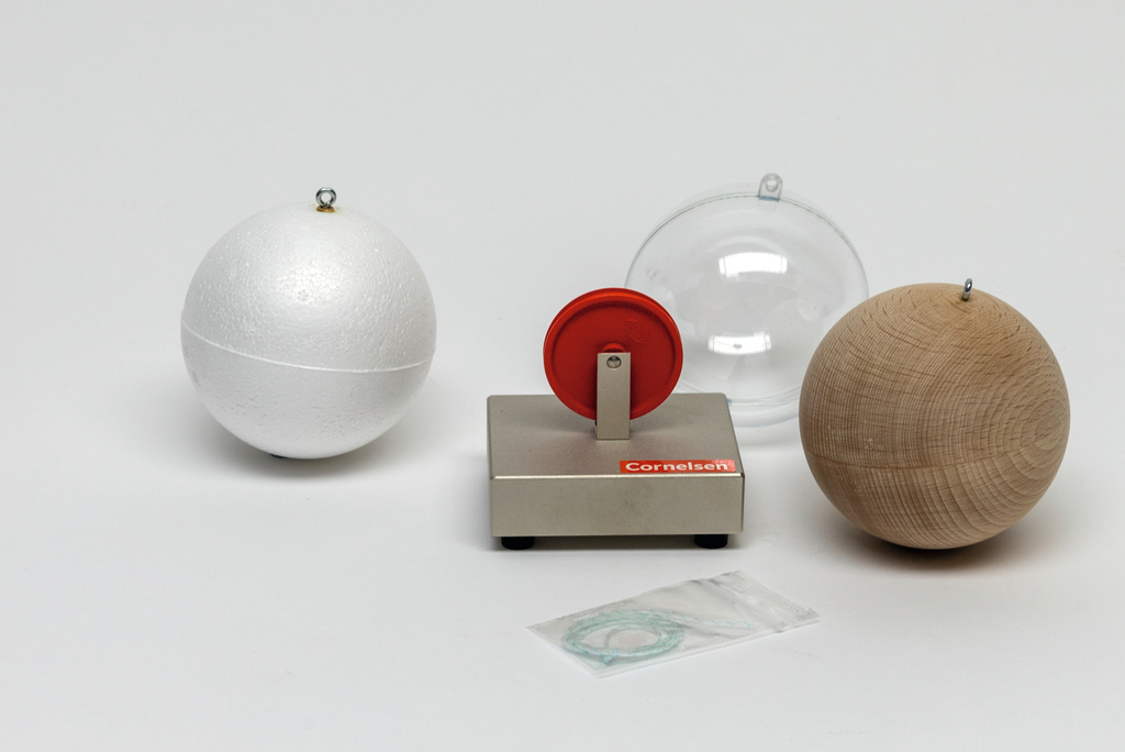 Das Bild zeigt eine Experimentierstation mit drei Kugeln aus verschiedenen Materialien: eine weiße Styroporkugel, eine Holz- und eine Kunststoffkugel, sowie ein rot-weißer Drehmechanismus. Dazu liegt ein kleines Paket mit einem Faden bereit, wahrscheinlich für den Gebrauch im Experiment.