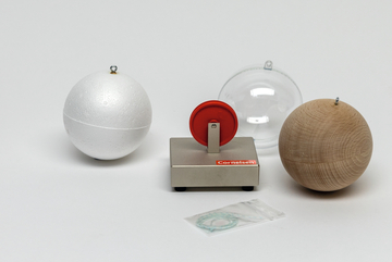 Das Bild zeigt eine Experimentierstation mit drei Kugeln aus verschiedenen Materialien: eine weiße Styroporkugel, eine Holz- und eine Kunststoffkugel, sowie ein rot-weißer Drehmechanismus. Dazu liegt ein kleines Paket mit einem Faden bereit, wahrscheinlich für den Gebrauch im Experiment.