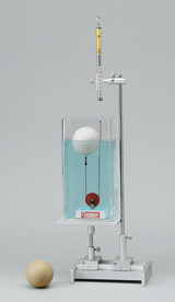 Das Bild zeigt ein Experimentiergerät mit einer transparenten Wanne, die mit blauem Wasser gefüllt ist, in der eine weiße Kugel schwimmt. Oben hängt eine Dropleiste, die zur Messung von Kräften oder Dichten verwendet wird.