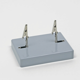 Das Bild zeigt eine kleine graue Basis mit zwei Krokodilklemmen, die an der Oberseite befestigt sind. Diese Vorrichtung wird häufig in Experimenten zur Durchführung elektrischer Schaltungen oder zum Halten von Drähten verwendet.