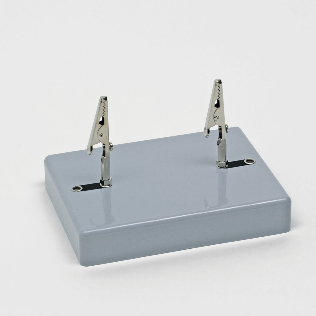 Das Bild zeigt eine kleine graue Basis mit zwei Krokodilklemmen, die an der Oberseite befestigt sind. Diese Vorrichtung wird häufig in Experimenten zur Durchführung elektrischer Schaltungen oder zum Halten von Drähten verwendet.