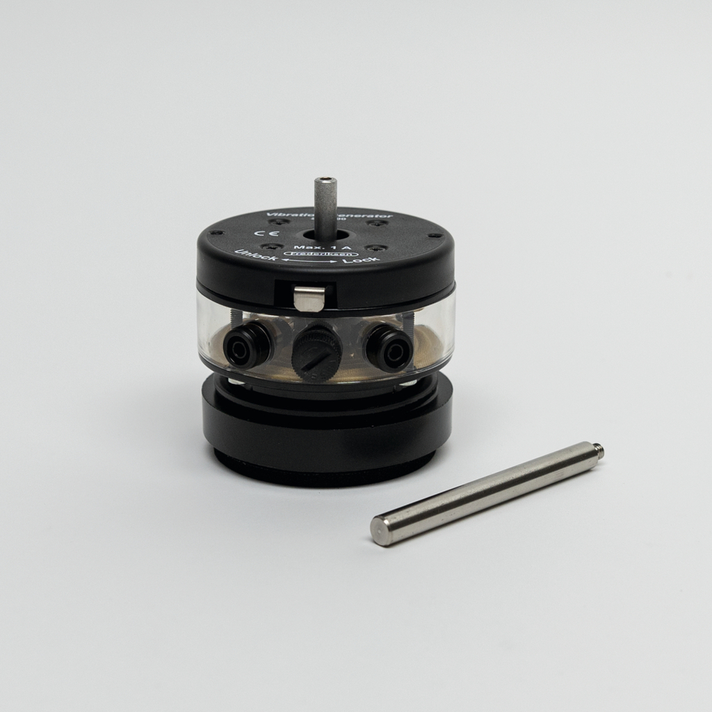 Das Bild zeigt einen Vibrationsgenerator mit einem transparenten Gehäuse und Steuerknöpfen. Neben dem Gerät liegt ein metallischer Stab, der möglicherweise als Zubehör dient.