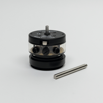 Das Bild zeigt einen Vibrationsgenerator mit einem transparenten Gehäuse und Steuerknöpfen. Neben dem Gerät liegt ein metallischer Stab, der möglicherweise als Zubehör dient.