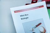 Das Bild zeigt die Gebrauchsanleitung einer Mini-Box für biologischen Experimentierbedarf im deutschen Bildungswesen. Auf der Anleitung ist ein Bild zu sehen, das eine Essenzubereitung mit einem Messer und einem Apfel zeigt.