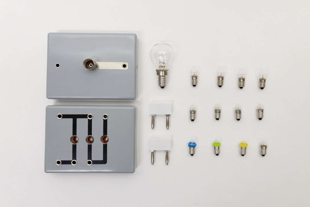 Das Bild zeigt ein Experimentier-Set mit zwei grauen Bauteilen, die mit elektrischen Schaltungen verbunden sind, sowie verschiedenen Glühbirnen in unterschiedlichen Farben. Es beinhaltet außerdem zwei Stecker, die für die elektrischen Verbindungen verwendet werden können.