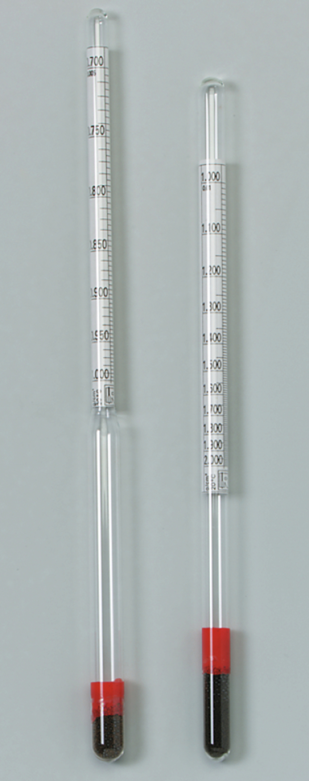 Das Bild zeigt zwei sichtbare Messzylinder aus Glas mit roten Kunststoffstopfen am Boden, die Skalen zur Abmessung von Flüssigkeiten oder Feststoffen aufweisen. Diese Geräte sind typisches Experimentiermaterial für den naturwissenschaftlichen Unterricht.