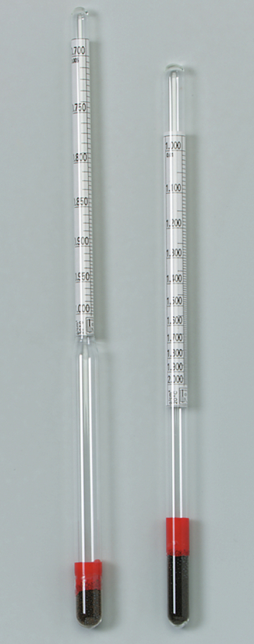 Das Bild zeigt zwei sichtbare Messzylinder aus Glas mit roten Kunststoffstopfen am Boden, die Skalen zur Abmessung von Flüssigkeiten oder Feststoffen aufweisen. Diese Geräte sind typisches Experimentiermaterial für den naturwissenschaftlichen Unterricht.
