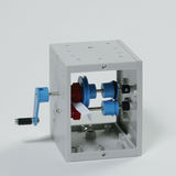 Das Bild zeigt ein Experimentiergerät in einem offenen Gehäuse, mit zwei rotierenden Elementen und einem Handgriff zur Betätigung. Es ist wahrscheinlich für lehrreiche Zwecke im physikalischen Unterricht konzipiert, um Konzepte wie Mechanik oder Energieübertragung zu veranschaulichen.