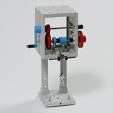 Das Bild zeigt eine mechanische Konstruktion mit einem offenen Gehäuse, in dem sich Zahnräder in roten und blauen Farben befinden. Diese Apparatur könnte zur Demonstration von Mechanik und Kraftübertragung im Bildungsbereich verwendet werden.