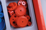 Das Bild zeigt eine Sammlung von roten Zahnrädern aus Kunststoff, die in einer Box organisiert sind. Diese Teile eignen sich für experimentelle Anwendungen im Bildungsbereich, um mechanische Konzepte zu veranschaulichen.