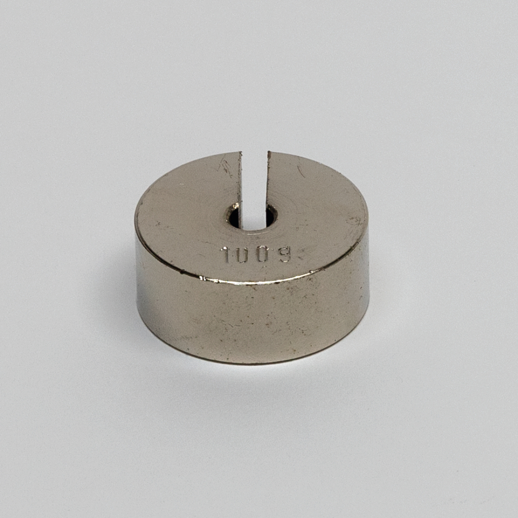 Das Bild zeigt einen runden, metallischen Block mit einer Vertiefung und einer Spalte in der Mitte, auf dem die Zahl "100 g" eingraviert ist. Dieses Objekt dient vermutlich als Gewicht für Experimente im Bildungswesen.