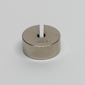 Das Bild zeigt einen runden, metallischen Block mit einer Vertiefung und einer Spalte in der Mitte, auf dem die Zahl "100 g" eingraviert ist. Dieses Objekt dient vermutlich als Gewicht für Experimente im Bildungswesen.