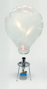 Das Bild zeigt einen transparenten, ballonartigen Behälter, der über einem kleinen Campingkocher platziert ist. Der Kocher enthält eine Gasflasche, die vermutlich für Experimente zur Dampf- oder Wärmeerzeugung verwendet wird.
