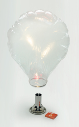 Das Bild zeigt ein transparentes, ballonähnliches Gefäß, das von einem Metallständer gehalten wird. Darunter befindet sich eine Wärmequelle, während daneben eine Schachtel mit Streichhölzern liegt, die für Experimente im Bildungsbereich genutzt wird.