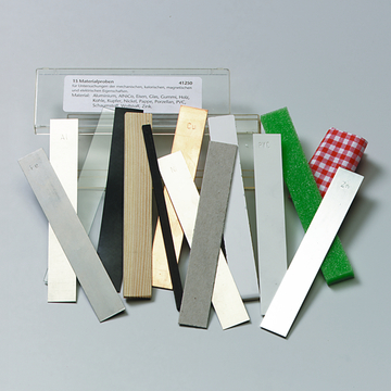 Das Bild zeigt eine Sammlung von 15 Materialproben, die für Experimente zu mechanischen, kalorischen und magnetischen Eigenschaften verwendet werden. Die Proben bestehen aus verschiedenen Materialien wie Aluminium, Eisen, Holz und PVC.