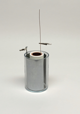 Das Bild zeigt ein Experimentiergerät, das aus einem gläsernen Zylinder mit einem metallischen Inneren und zwei Klemmen besteht. Ein Draht ist durch eine Öffnung im oberen Teil des Zylinders geführt, was auf eine mögliche Verwendung zur Demonstration elektrischer Phänomene hindeutet.