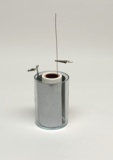 Das Bild zeigt ein Experimentiergerät, das aus einem gläsernen Zylinder mit einem metallischen Inneren und zwei Klemmen besteht. Ein Draht ist durch eine Öffnung im oberen Teil des Zylinders geführt, was auf eine mögliche Verwendung zur Demonstration elektrischer Phänomene hindeutet.