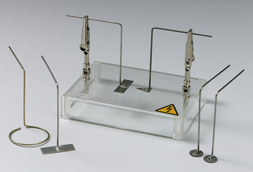 Das Bild zeigt ein Set von Experimentiergeräten, darunter Metallkontakte und eine transparente Basis mit einem Warnsymbol für Elektrizität. Dieses Equipment wird vermutlich für physikalische Experimente im schulischen Kontext verwendet.