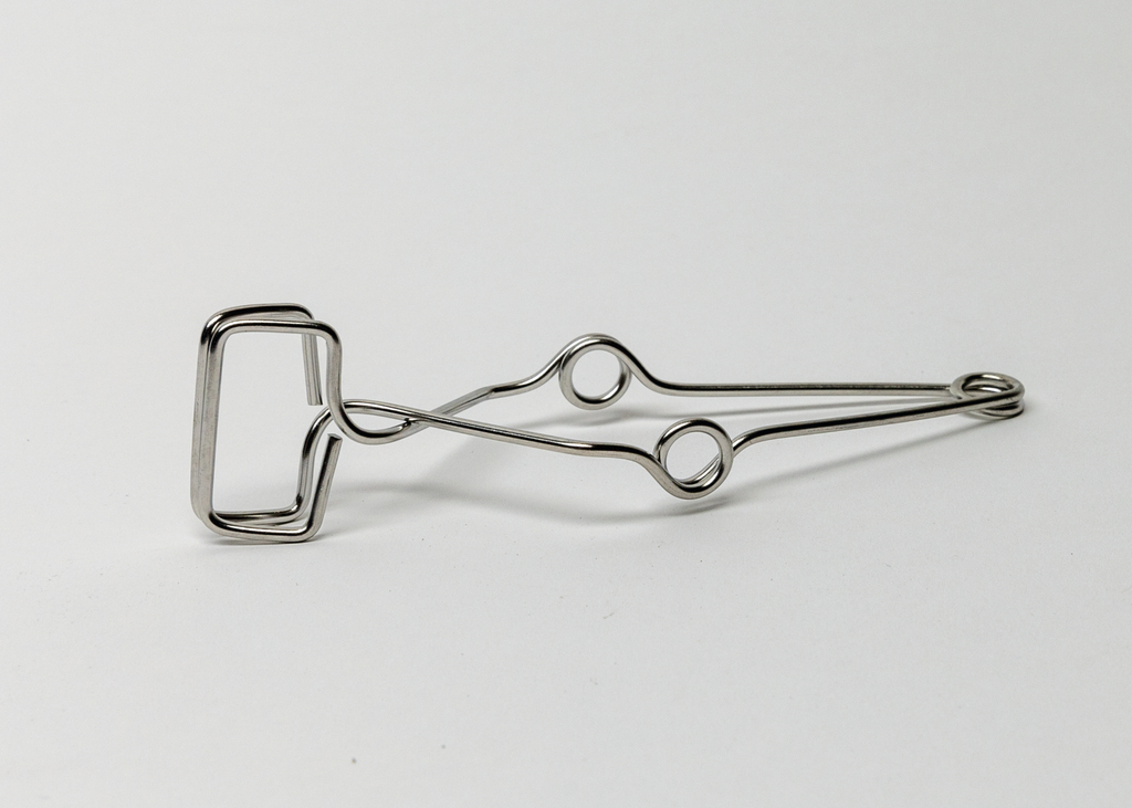 Das Bild zeigt eine metallene Klammer mit einem rechteckigen Greifmechanismus und zwei runden Öffnungen. Diese Klammer könnte für verschiedene Experimente im Bildungsbereich verwendet werden, um Materialien zu fixieren oder zu halten.