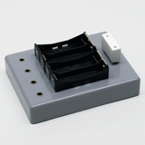 Das Bild zeigt eine graue Experimentierplatte mit zwei schwarzen Batteriehaltern und mehreren Anschlüssen. Sie ist für den Einsatz im Bildungsbereich konzipiert, um elektrische Experimente zu ermöglichen.