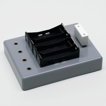 Das Bild zeigt eine graue Experimentierplatte mit zwei schwarzen Batteriehaltern und mehreren Anschlüssen. Sie ist für den Einsatz im Bildungsbereich konzipiert, um elektrische Experimente zu ermöglichen.