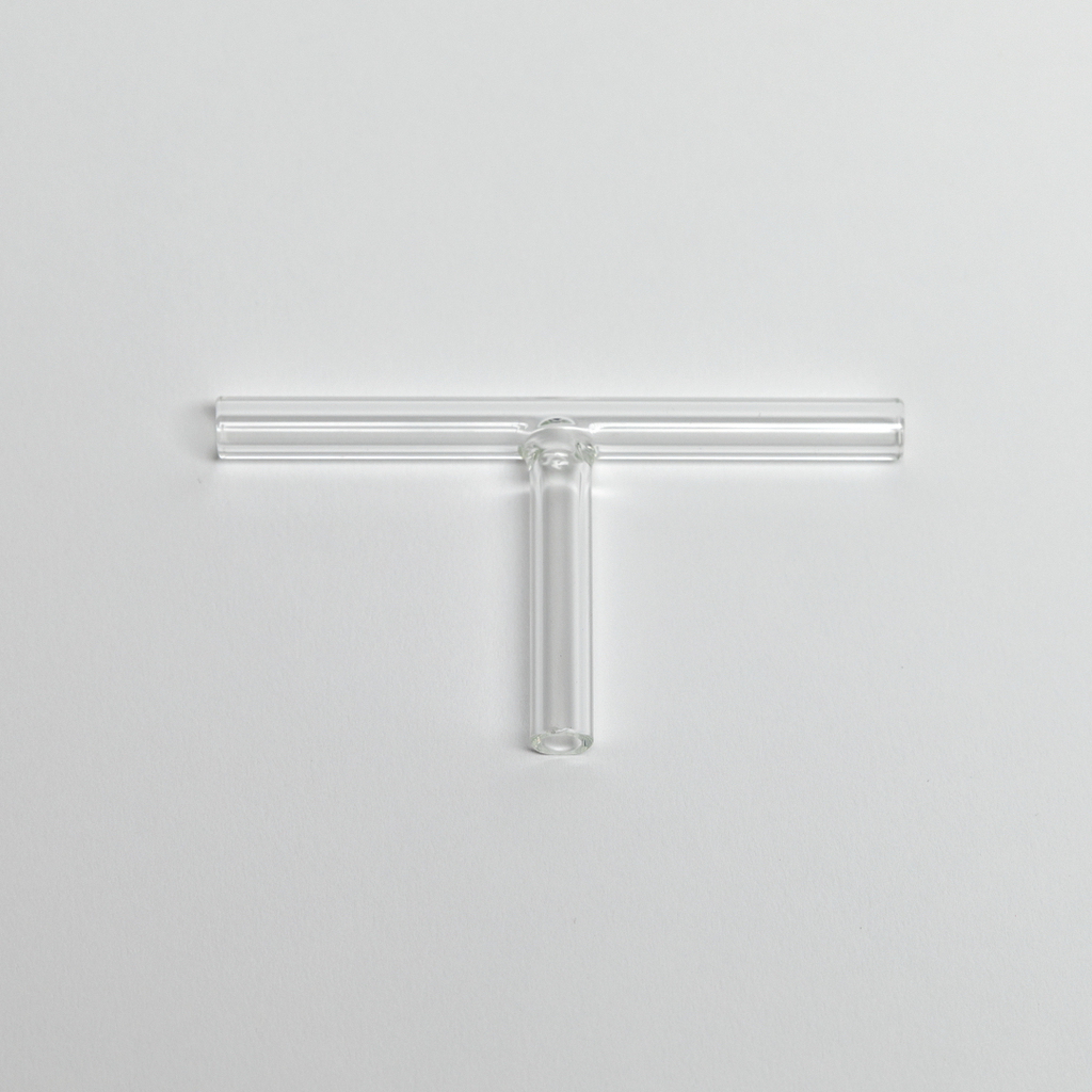 Das Bild zeigt ein transparentes Glasgerät in Form eines "T", das vermutlich aus einem Experimentierbedarf für chemische oder physikalische Versuche stammt. Es besteht aus mehreren Röhren, die in einem zentralen Teil verbunden sind, und dient wahrscheinlich zur Durchführung von Experimenten.