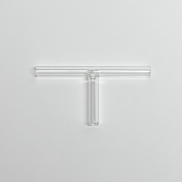 Das Bild zeigt ein transparentes Glasgerät in Form eines "T", das vermutlich aus einem Experimentierbedarf für chemische oder physikalische Versuche stammt. Es besteht aus mehreren Röhren, die in einem zentralen Teil verbunden sind, und dient wahrscheinlich zur Durchführung von Experimenten.