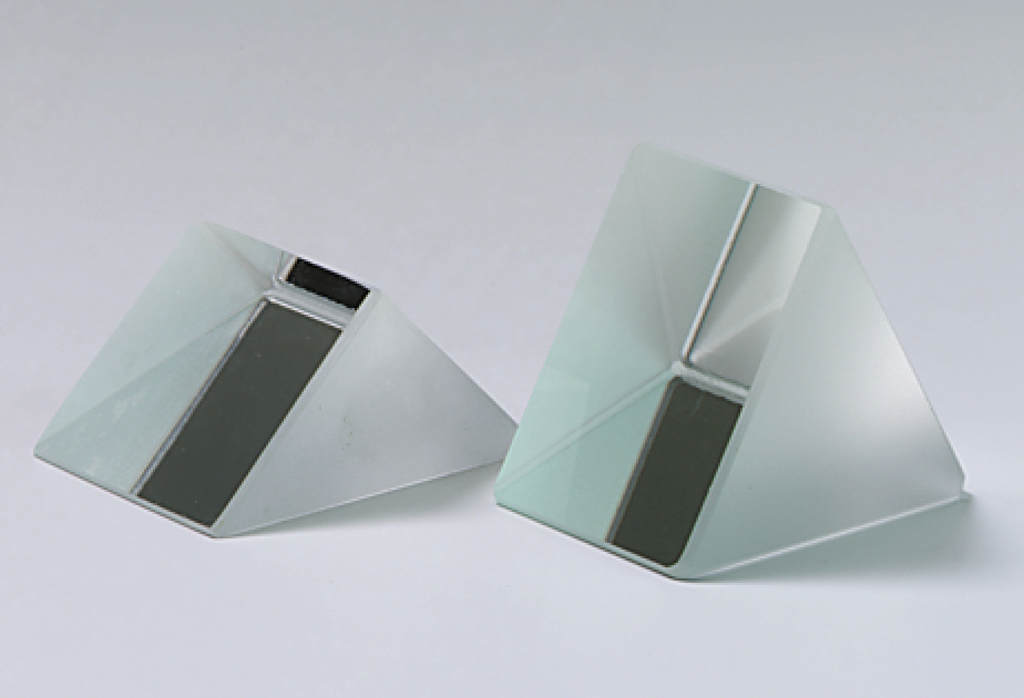 Das Bild zeigt zwei prismatische Glasobjekte, die leicht geneigt stehen. Sie sind jeweils halbdurchsichtig und haben eine flache, schwarze Fläche an der Seite, die für optische Experimente genutzt werden kann.