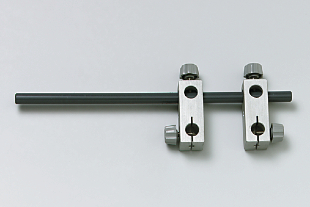 Das Bild zeigt ein Experimentiergerät mit einem grauen Stab und zwei metallischen Halterungen. Diese Halterungen ermöglichen die Befestigung des Stabs in unterschiedlichen Positionen, vermutlich für physikalische Experimente.