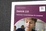Das Bild zeigt ein Buchcover mit dem Titel "Elektrik 2.0", das einen Klassensatz für Grundschaltungen auf einer Steckplatte vorstellt. Es enthält einen QR-Code zur Unterstützung und zeigt einen Jungen, der an einem Experiment arbeitet.