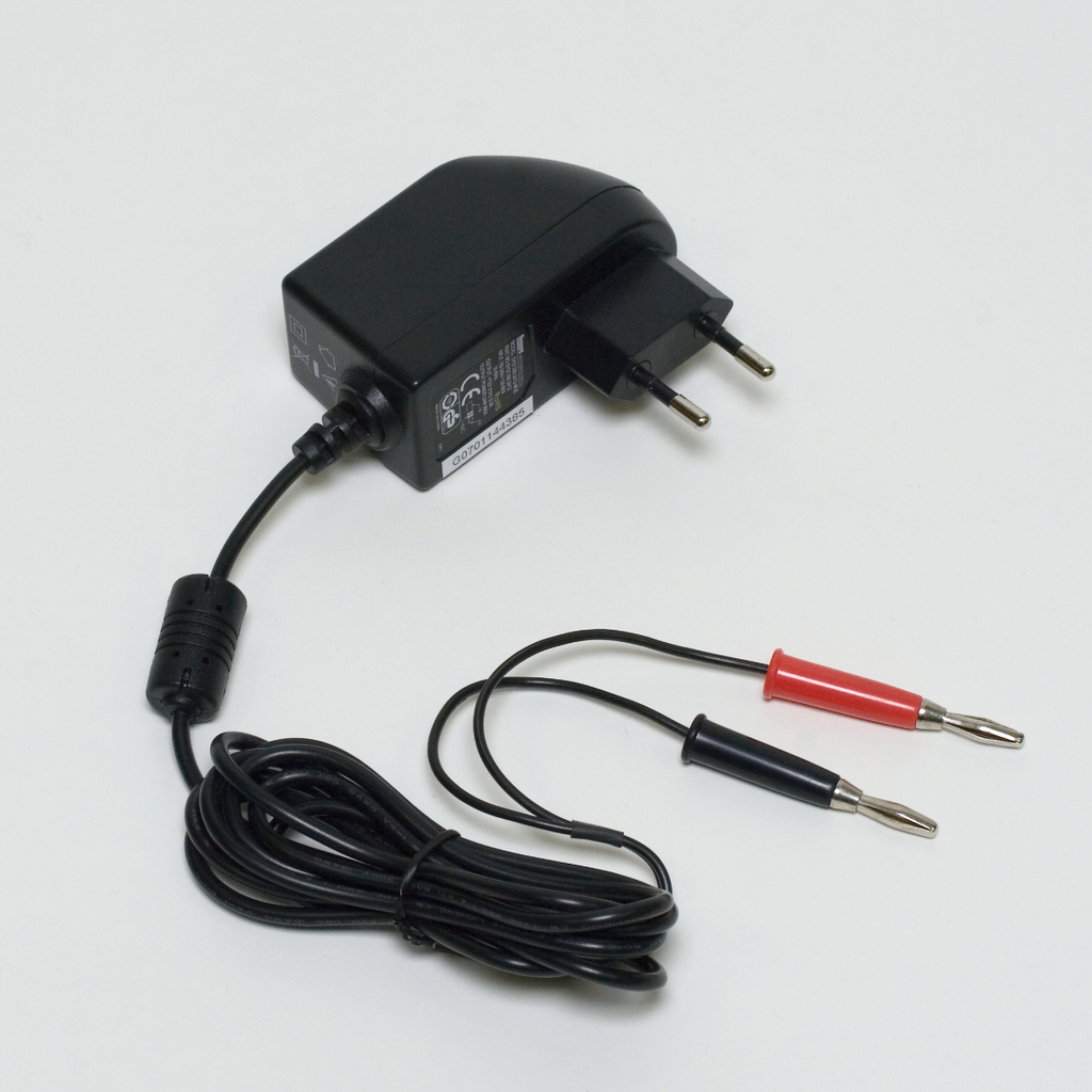 Das Bild zeigt ein schwarzes Netzteil mit einem Eurostecker und zwei Kabeln mit Krokodilklemmen in Rot und Schwarz. Es ist für den Einsatz in Experimenten im Bildungswesen konzipiert und stellt eine Verbindung für elektrische Anwendungen her.