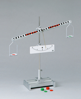 Das Bild zeigt eine Wippe mit zwei Schalen, die an einem horizontalen Balken angebracht sind, der an einem zentralen Drehpunkt gelagert ist. Zusätzlich liegen farbige Scheiben in rot und grün auf dem Tisch, die vermutlich zur Gewichtsermittlung oder zum Experimentieren verwendet werden.