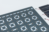 Das Bild zeigt eine experimentelle Platine mit mehreren quadratischen Kontakten und Leitungen, die in einem regelmäßig angeordneten Muster angebracht sind. Rechts sind kleine Solarmodule zu sehen, die für Experimente mit erneuerbarer Energie verwendet werden können.
