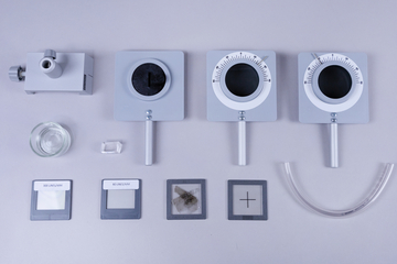 Das Bild zeigt eine Auswahl an Experimentiergeräten, darunter Messinstrumente wie eine Ablenkröhre und verschiedene Objektträger. Zusätzlich sind ein Glasbehälter und ein transparentes Rohr zu sehen, die für physikalische Experimente genutzt werden können.