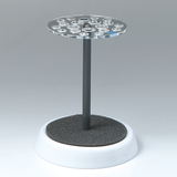 Das Bild zeigt ein Experimentiergerät, das aus einem runden, transparenten Oberteil mit mehreren Vertiefungen auf einem stabilen, schwarzen Ständer besteht. Es dient wahrscheinlich zur sicheren Aufbewahrung von Reagenzgläsern oder ähnlichem Laborgeschirr.