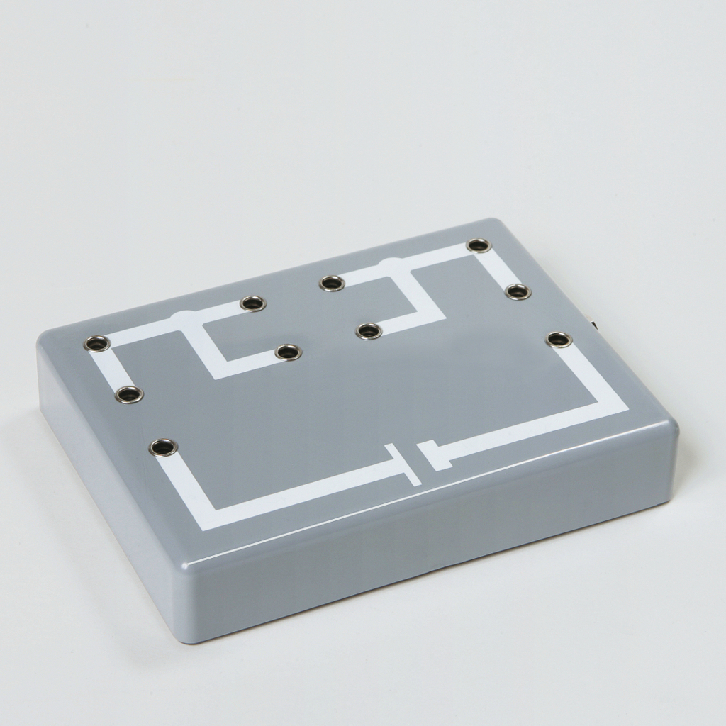 Das Bild zeigt eine graue Experimentierplatte mit weißen Linien, die ein Schaltplan-Diagramm darstellt. Die Oberfläche weist mehrere Anschlussbuchsen auf, die für elektrische Experimente verwendet werden können.
