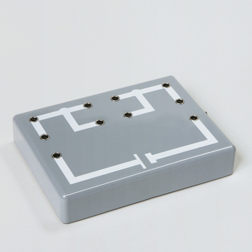 Das Bild zeigt eine graue Experimentierplatte mit weißen Linien, die ein Schaltplan-Diagramm darstellt. Die Oberfläche weist mehrere Anschlussbuchsen auf, die für elektrische Experimente verwendet werden können.