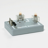 Das Bild zeigt eine Experimentier-Station bestehend aus einem grauen Gehäuse mit zwei Glühbirnen und einem Kippschalter. Diese Vorrichtung dient zur Demonstration von elektrischen Schaltkreisen im Bildungswesen.