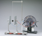 Das Bild zeigt ein physikalisches Experimentiergerät bestehend aus einer Pendelkugel, die zwischen zwei Metallscheiben hängt, und einem rotierenden Rad mit elektrischen Kontakten. Es dient zur Demonstration von Kräften und Energieübertragung in physikalischen Experimenten im Bildungsbereich.