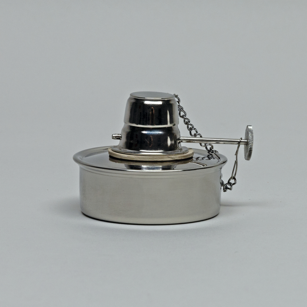 Das Bild zeigt einen Edelstahl-Brenner, der typischerweise in labortechnischen Experimenten verwendet wird. Er besitzt einen mittigen Docht und eine Schutzkappe, die mit einer Kette verbunden ist.