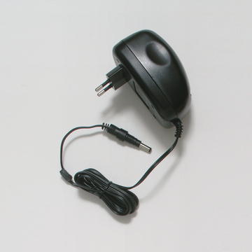 Das Bild zeigt ein schwarzes Netzteil mit einem Eurostecker, das für die Stromversorgung elektrischer Geräte verwendet wird. Es hat ein abgehendes Kabel mit einem Rundstecker für den Anschluss an verschiedene Geräte oder Experimentierbedarf.