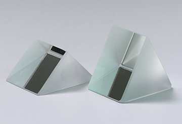 Das Bild zeigt zwei prismatische Glasskörper mit einer matten Oberfläche und einer horizontalen, schwarzen Fläche. Diese geometrischen Objekte werden häufig für physikalische Experimente verwendet, um Lichtbrechung und Reflexion zu demonstrieren.