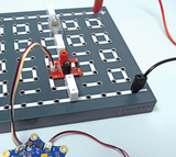 Das Bild zeigt eine Experimentierplatte mit elektrischen Komponenten, darunter ein Modul mit Anschlüssen und einer Batterie. Flexible Anschlussdrähte ermöglichen verschiedene elektrische Verbindungen für experimentelle Zwecke im Bildungswesen.